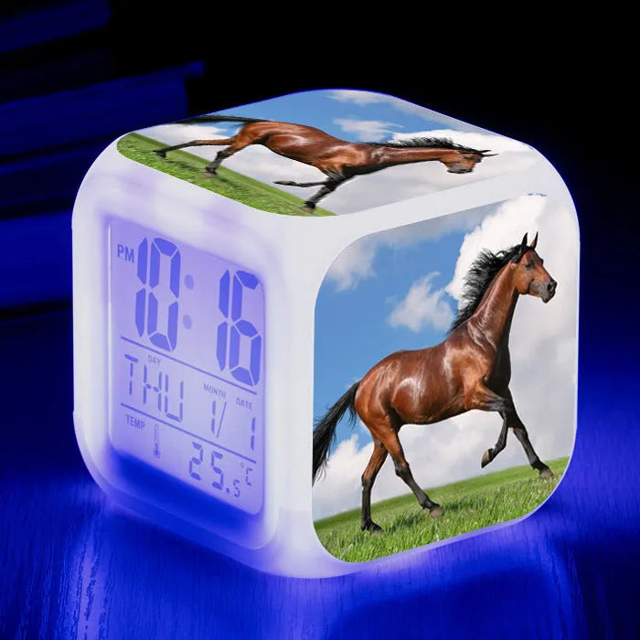 Square cartoon-style LED alarm clock showing colour-changing display on a bedside table.