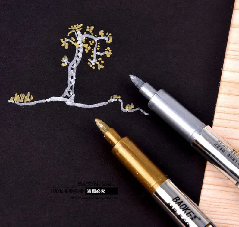 Gold and silver metallic marker pens on a glass surface showing fine tip ink marks