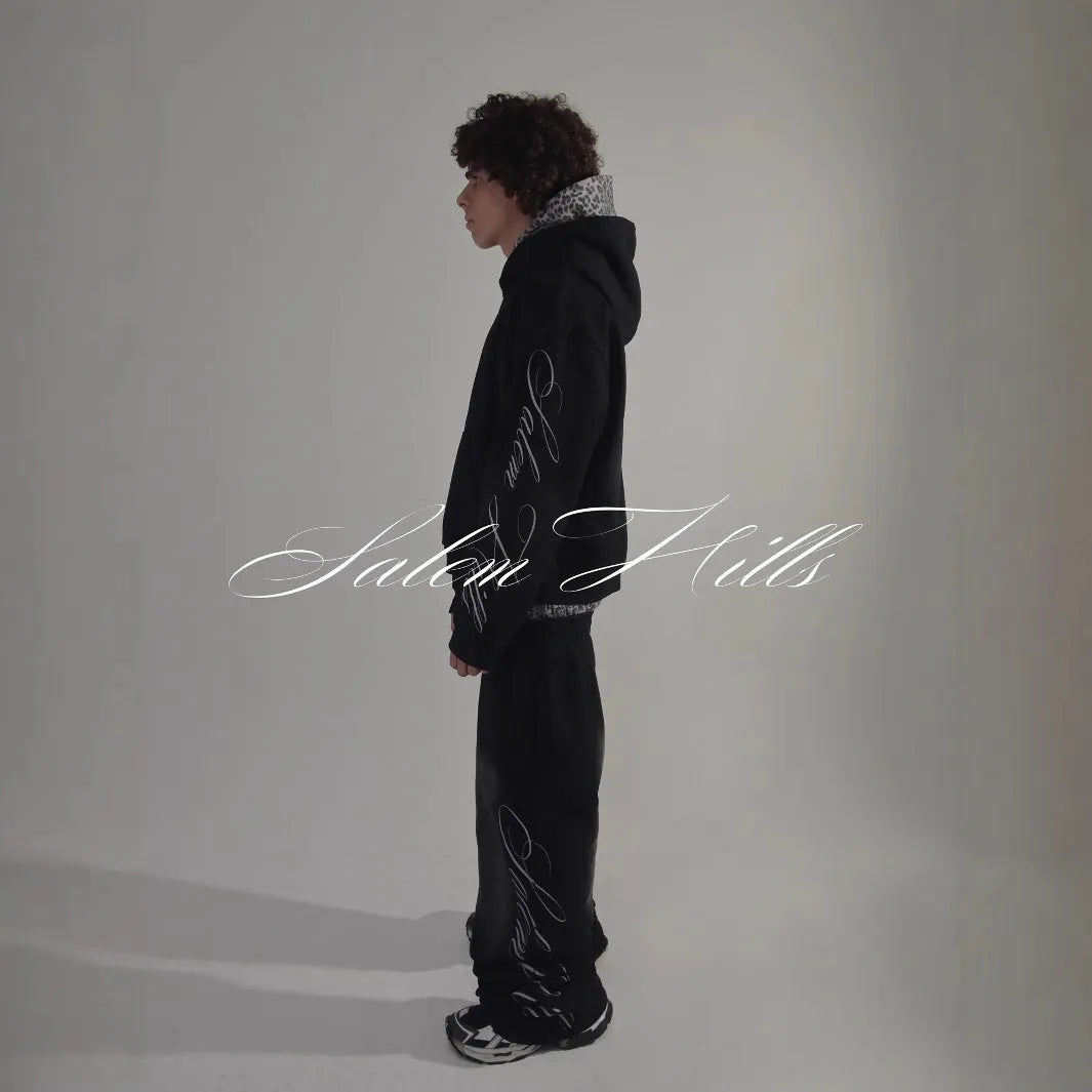 Man wearing a black punk-print hooded pullover jumpsuit with long sleeves and fleece lining.