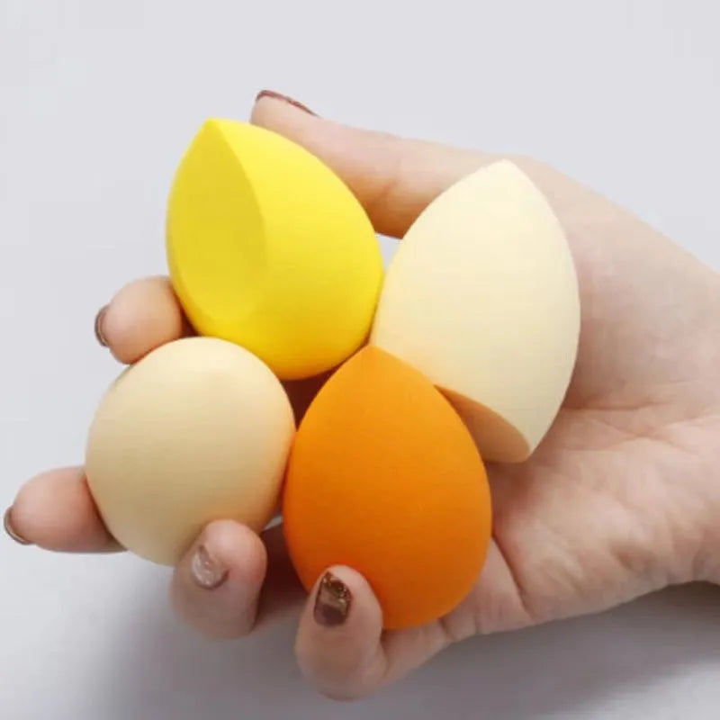 Set of seven differently shaped coloured makeup sponges including pear, heart, and drop shapes on a white background.