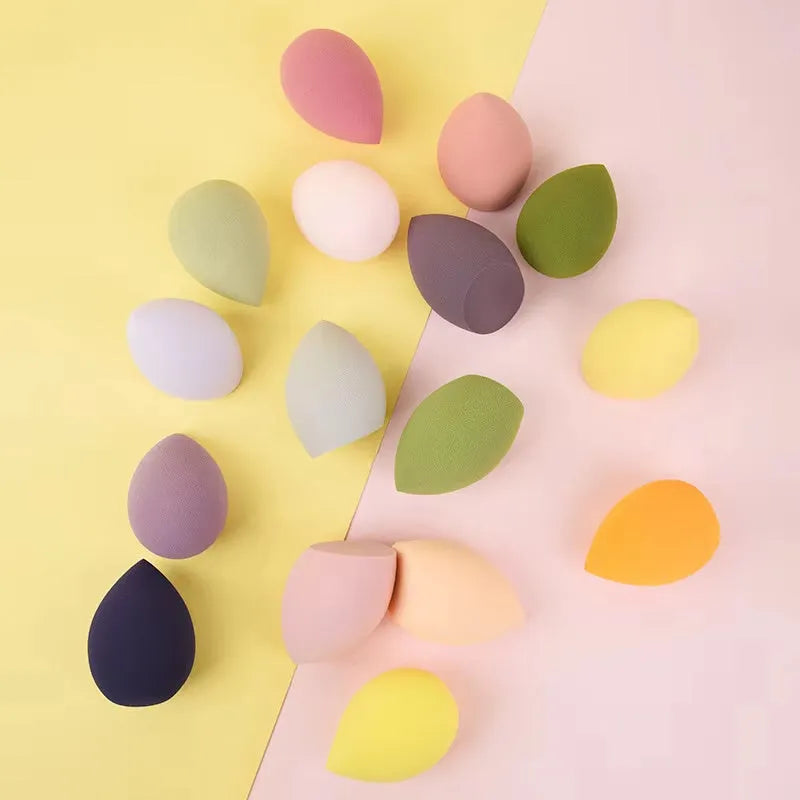 Set of seven differently shaped coloured makeup sponges including pear, heart, and drop shapes on a white background.