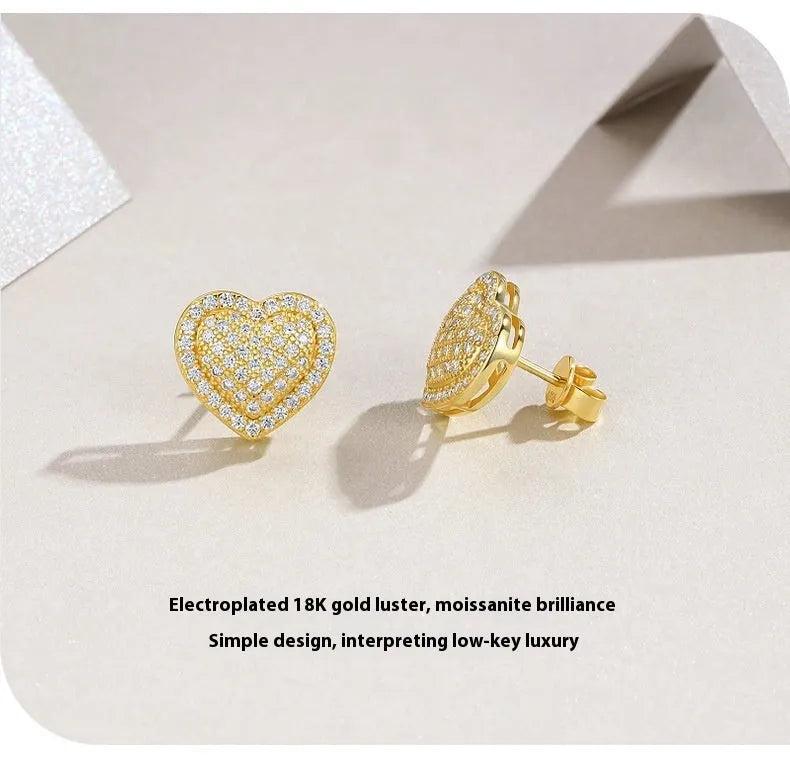 A pair of golden heart-shaped stud earrings with moissanite stones on a grey background.