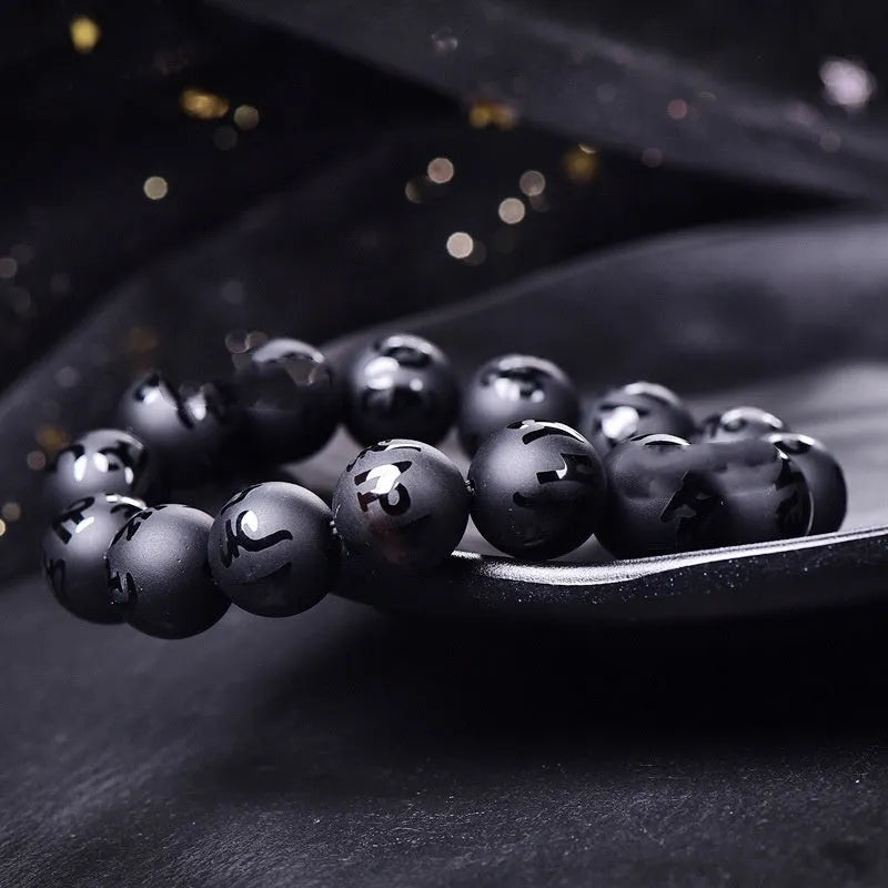 Close-up of a frosted black agate bracelet showing engraved Sanskrit mantra and Chinese-style patterns on a stone bead.