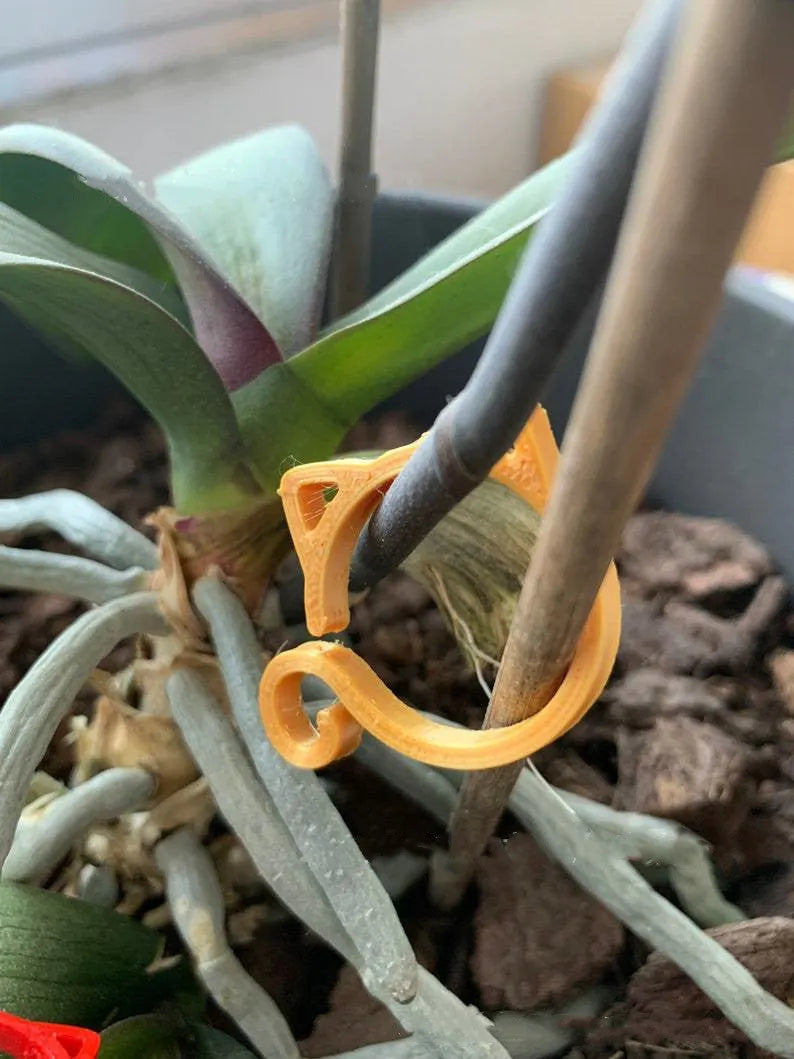 Red plastic cat-shaped clip for holding plant stems, shown against a green leaf background.