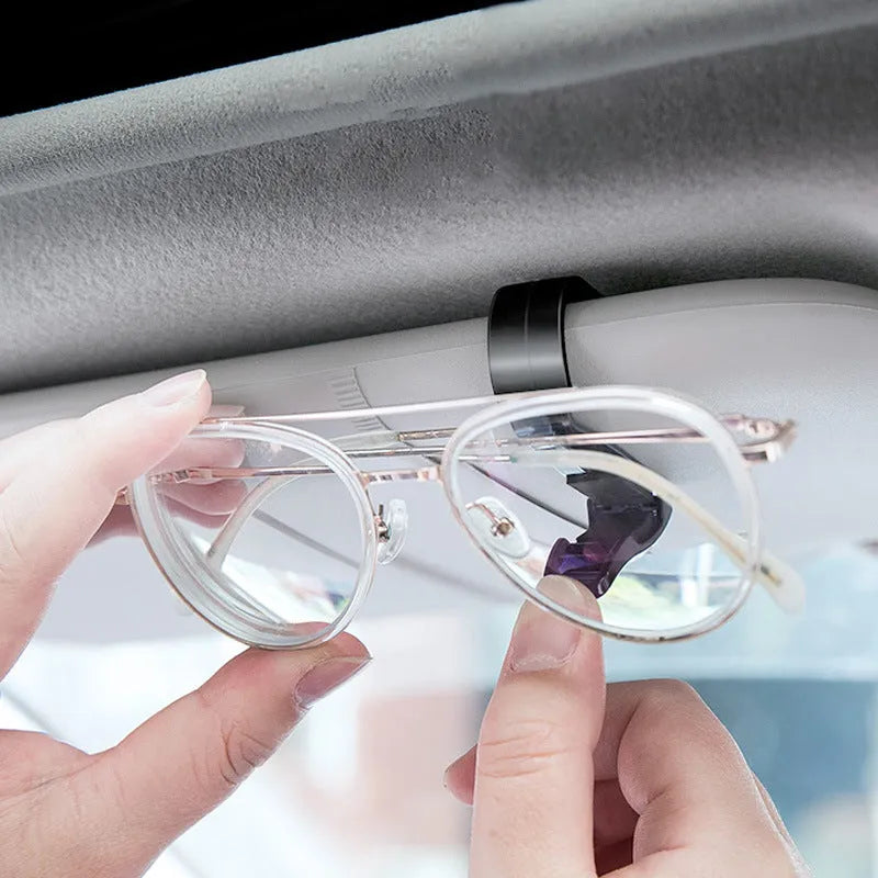 Black plastic clip mounted on a car sun visor holding a pair of sunglasses and an ID card.